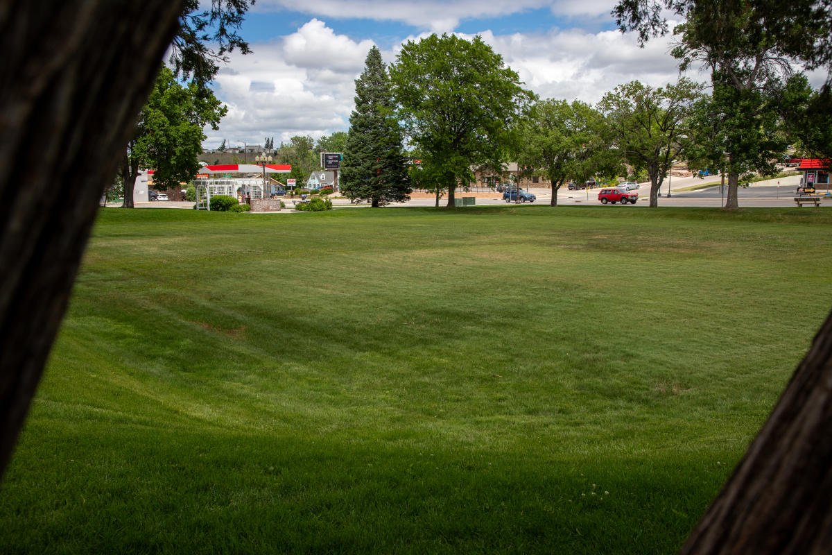 Wide open lawn at Wilson Park in Rapid City framed by mature shade trees. A white gazebo sits near the edge of the park, while nearby streets, local businesses, and neighborhood scenery create an inviting urban green space beneath a sky filled with puffy clouds.