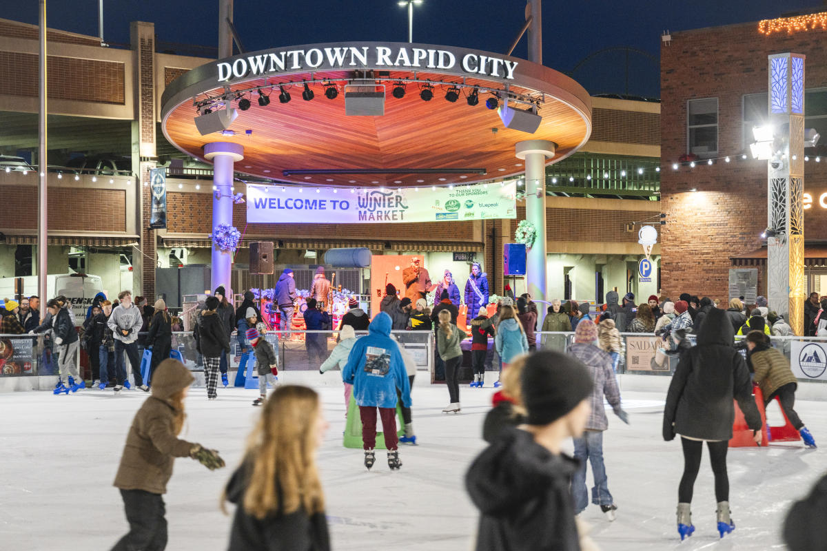 People skate on an ice rink in a lively winter market scene at night. A band performs on stage under a “Downtown Rapid City” sign, creating a festive atmosphere.