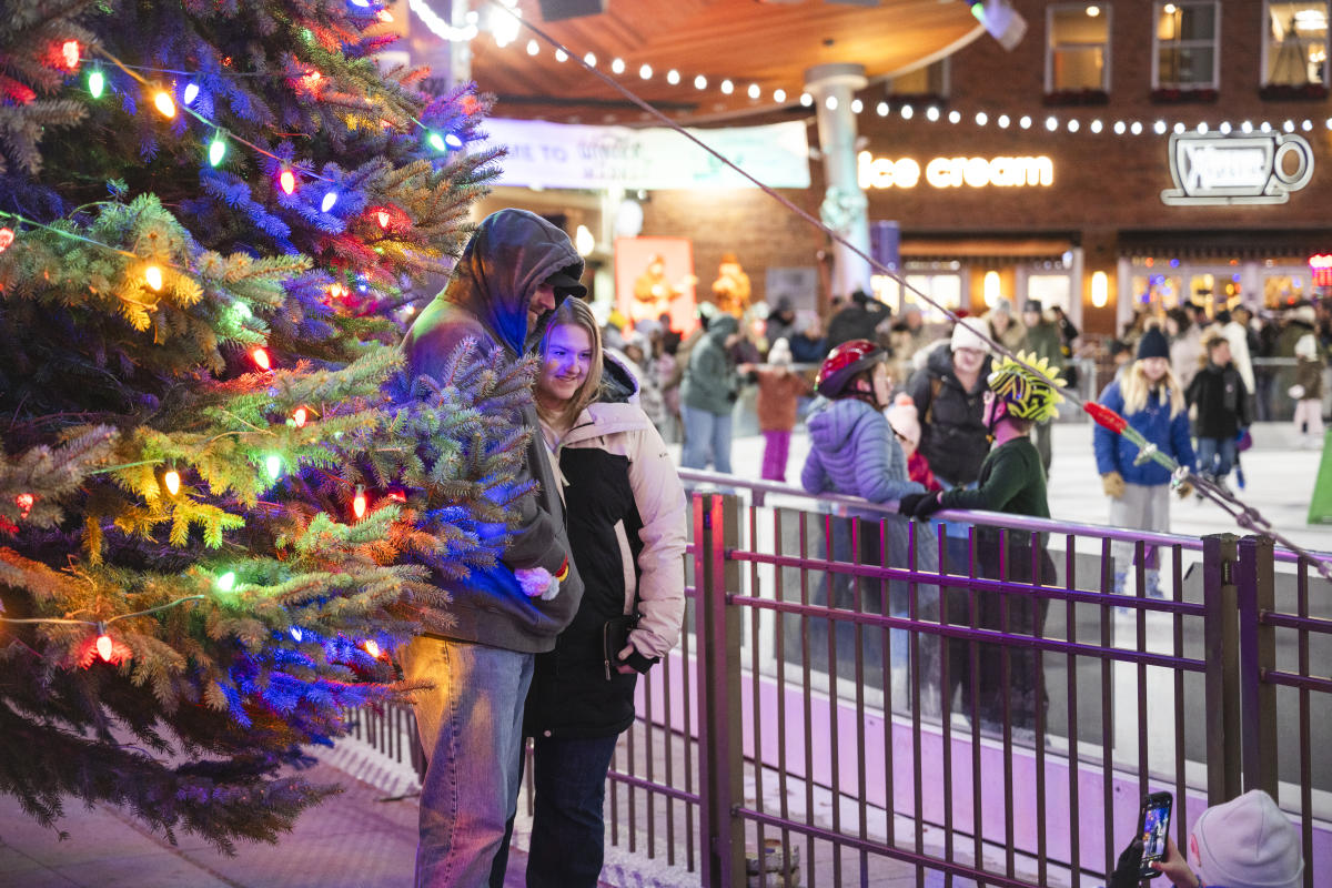 A couple stands warmly by a brightly lit holiday tree, surrounded by festive ice skaters. The scene is lively and joyful, evoking holiday cheer.