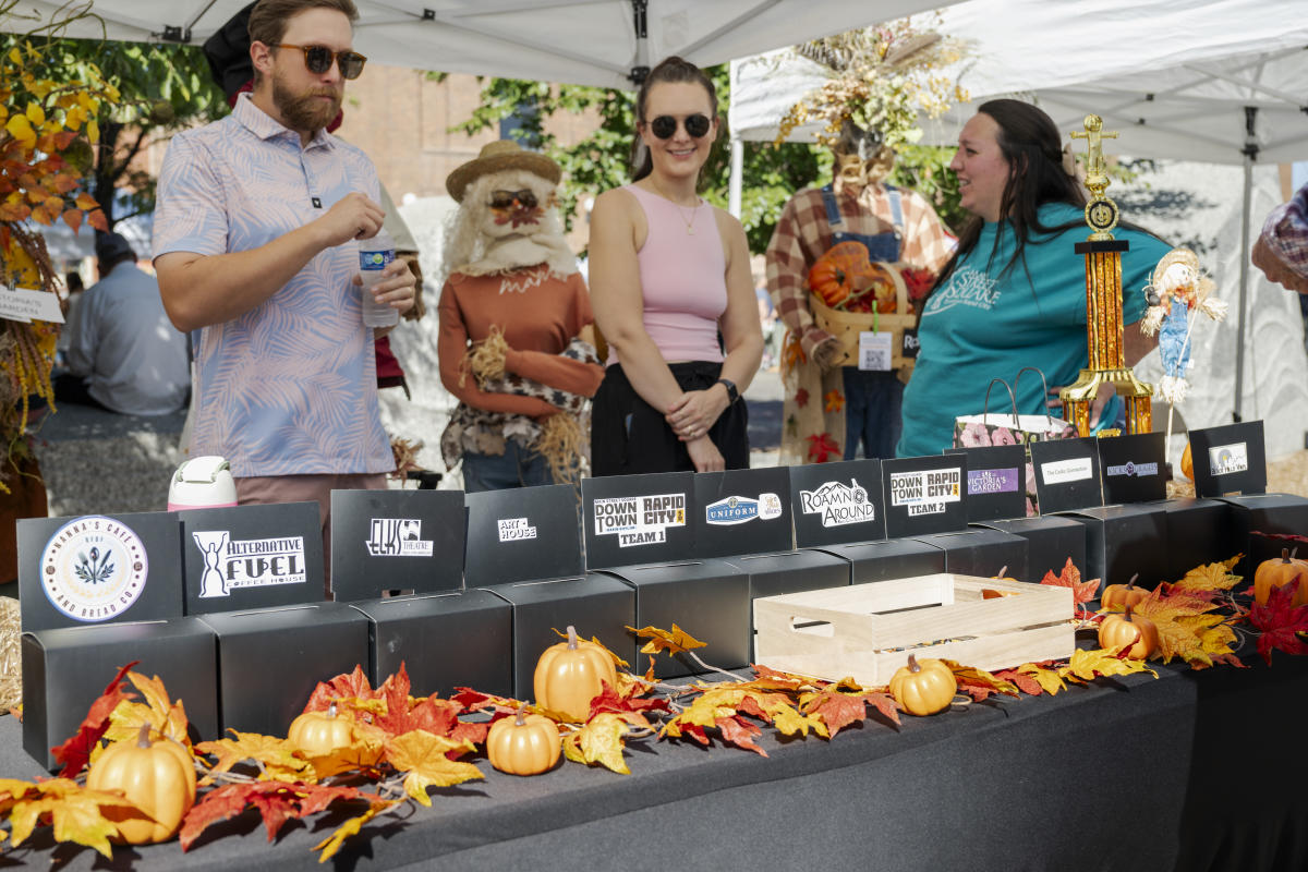 A festive market scene with three people under a tent. The table is decorated with autumn leaves and mini pumpkins, with boxes for different businesses for visitors to cast votes in.