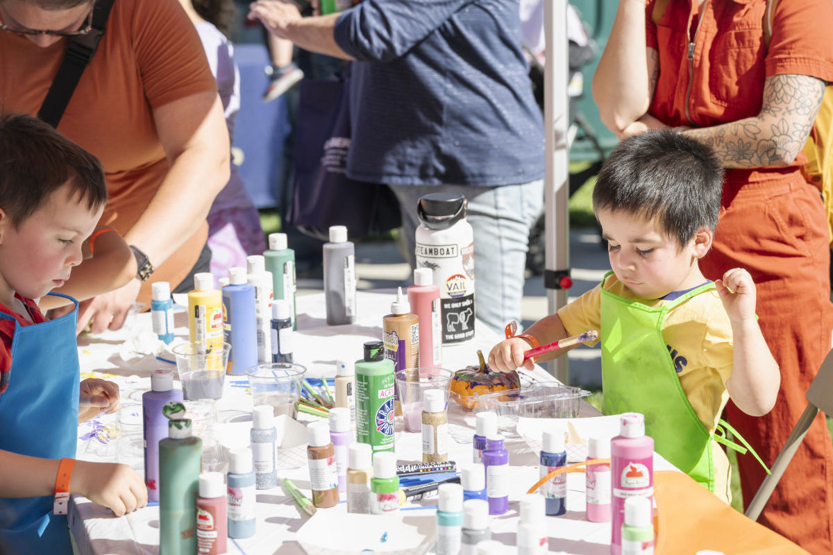 Children eagerly paint at a table filled with colorful paint bottles. The scene is lively and creative, with attentive adults nearby, and sunlight enhancing the playful mood.