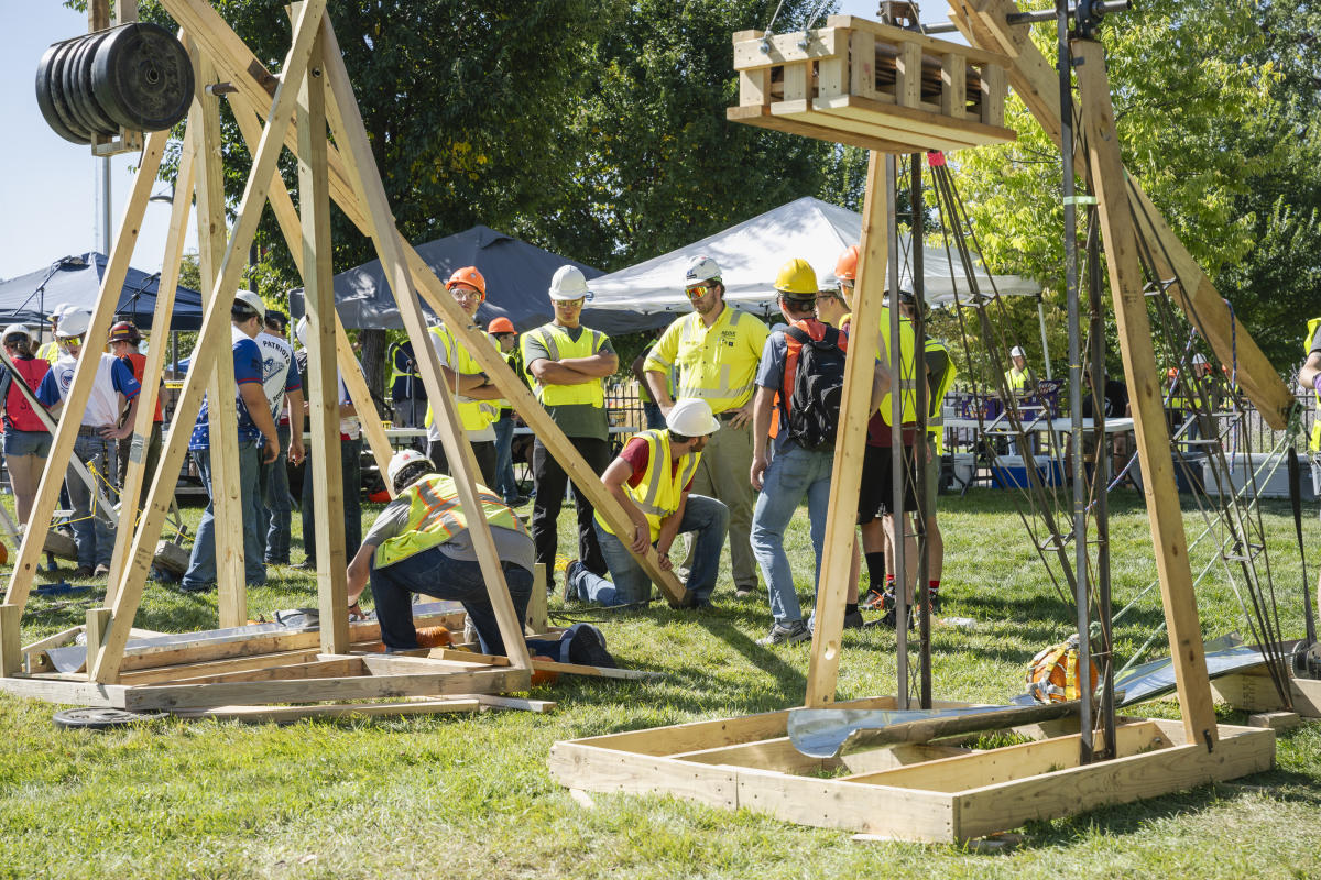 A group of SD Mines students in hard hats and vests gather around wooden trebuchets on a grassy field. The scene is busy and collaborative, with a sunny outdoor atmosphere.