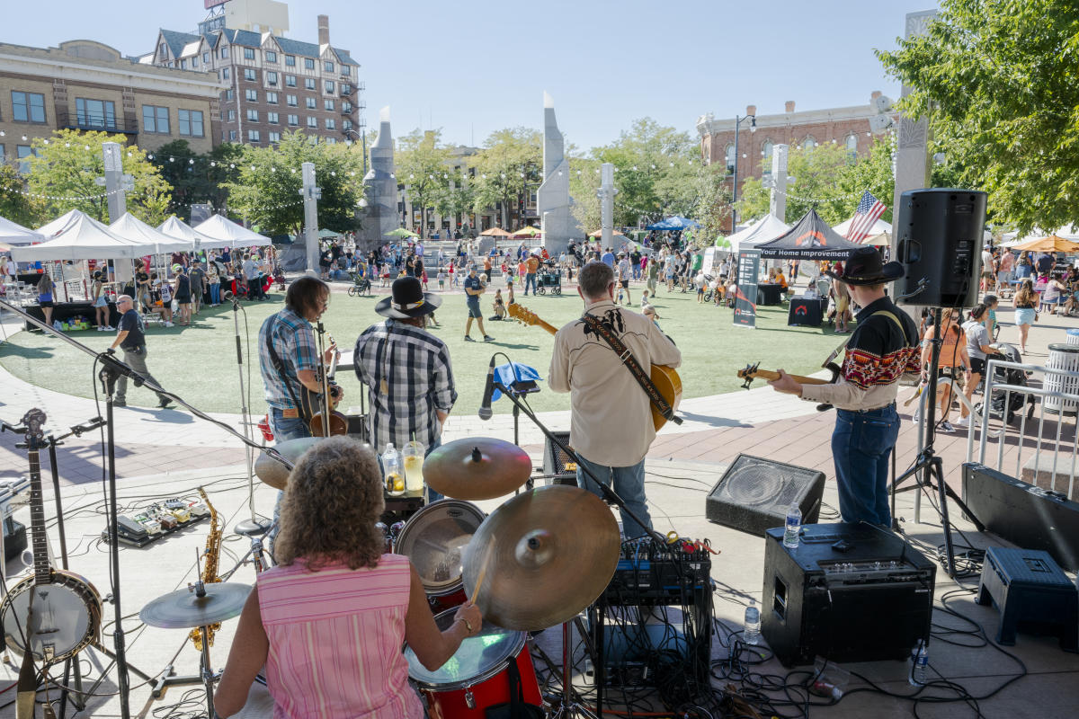 Wilt Brothers Band plays lively music on a stage at a the Downtown Pumpkin Festival. People browse vendor tents around a grassy area. The scene is vibrant and cheerful.