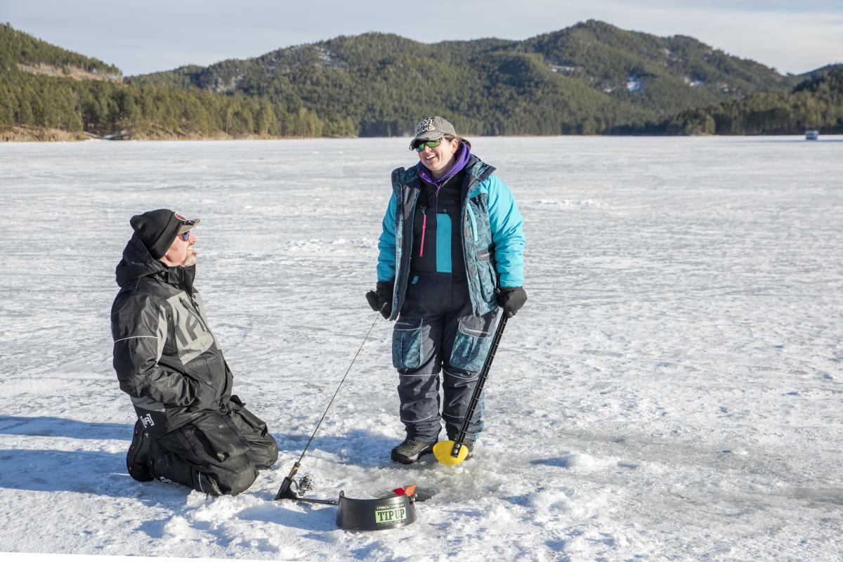 Two people in winter gear ice fish on a frozen lake, surrounded by snow and distant forested hills. The scene is bright and cheerful.