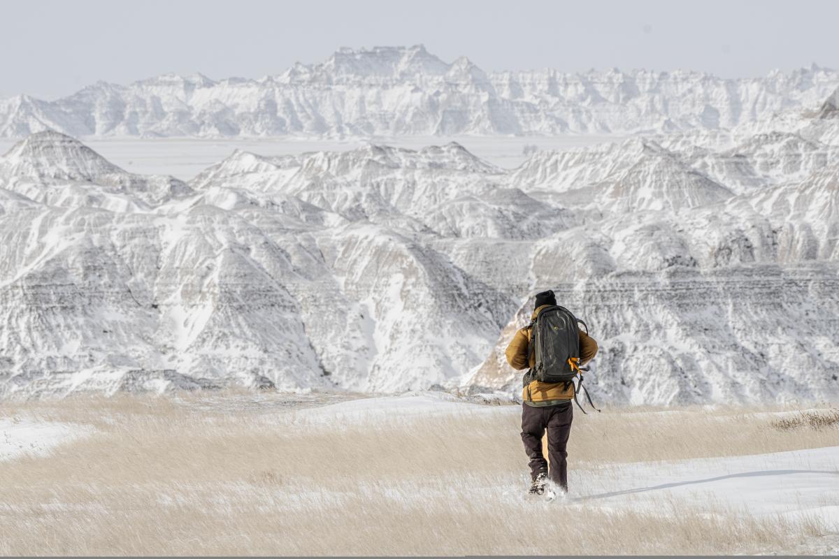 A person wearing winter clothing and a backpack hikes through snowy grasslands with snow-covered hills in the distance under a cloudy sky.