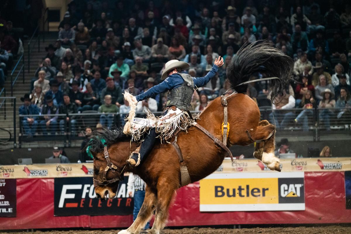 Rodeo scene with a cowboy in mid-air atop a bucking bronco, hand raised. The crowd watches from bleachers with banners and sponsors visible. Exciting and dynamic atmosphere.