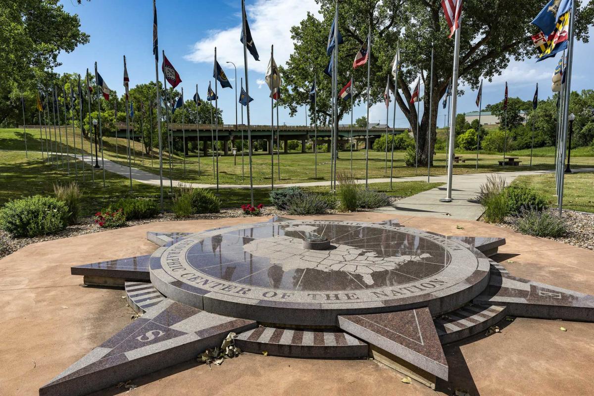 A stone monument in the shape of a compass that features a map of the United States is surrounded by state flags and mature trees.