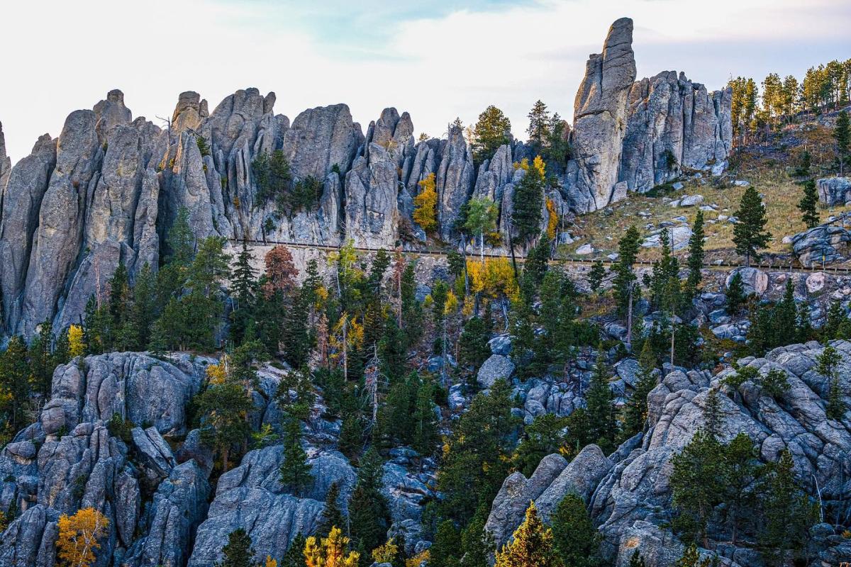 Jagged granite spires tower over a forested landscape with scattered autumn foliage. The scene is serene and dramatic under a partly cloudy sky.