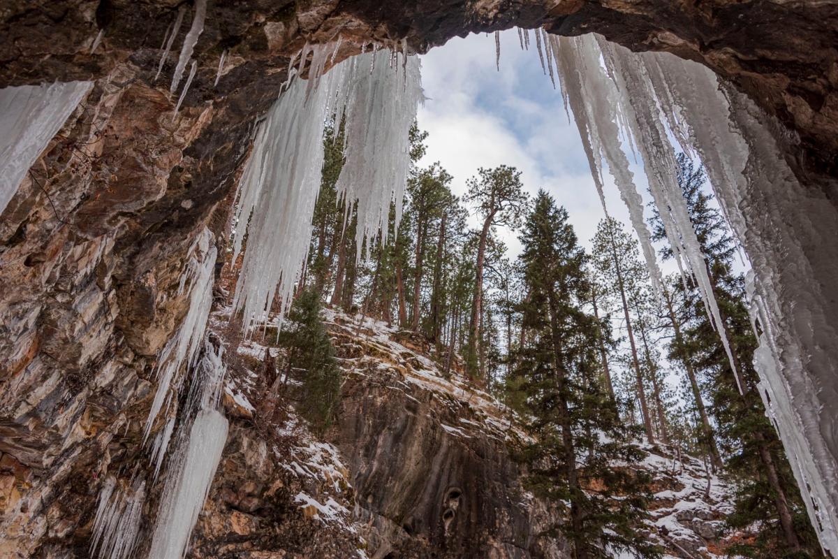 View from inside a rocky ice cave with large icicles hanging from the ceiling. Snow-dusted evergreen trees are visible outside against a cloudy sky.