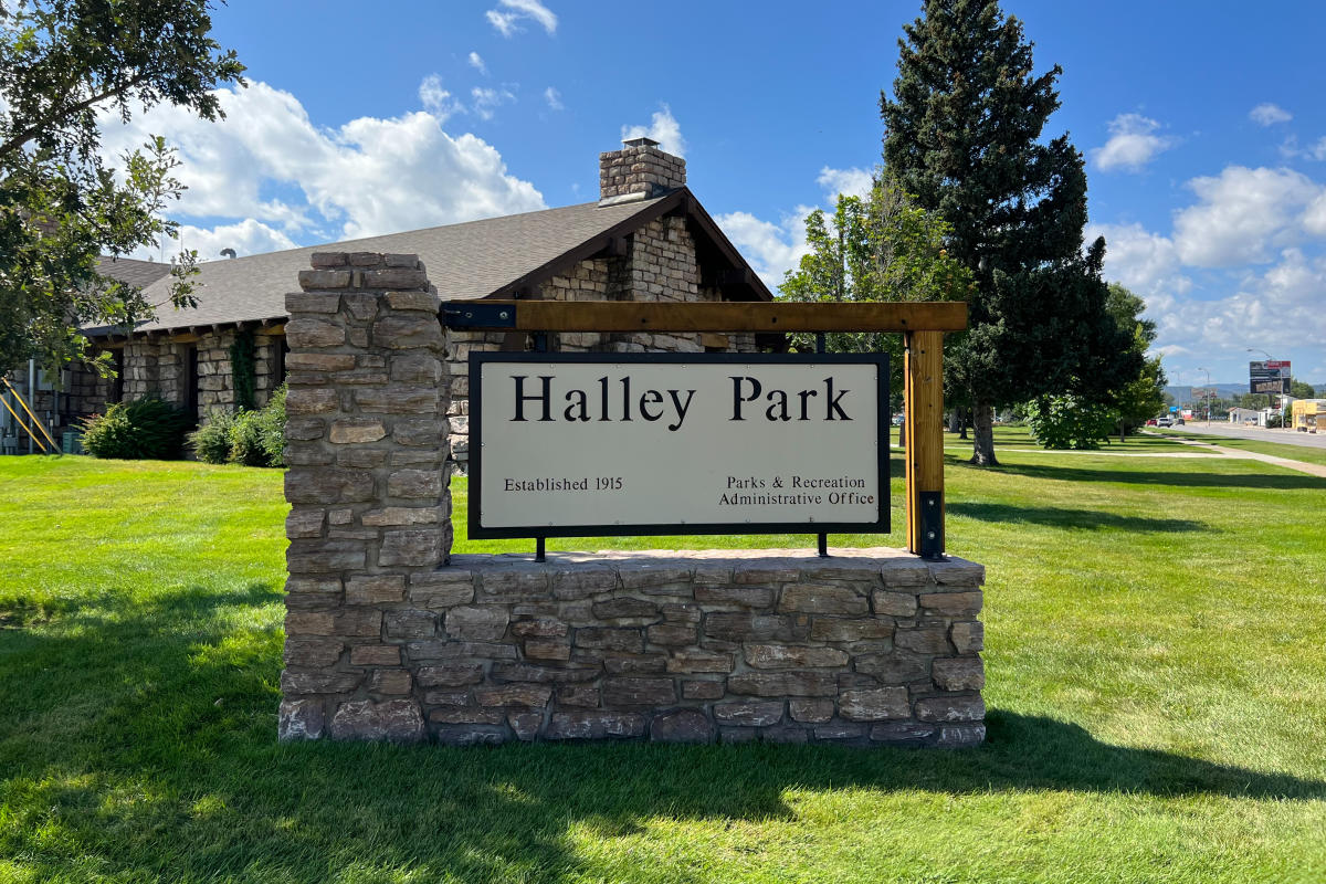 Stone monument sign for Halley Park in Rapid City stands on a manicured lawn outside the Parks & Recreation administrative office. Mature trees, open green space, and a bright blue sky with scattered clouds frame the historic park entrance.