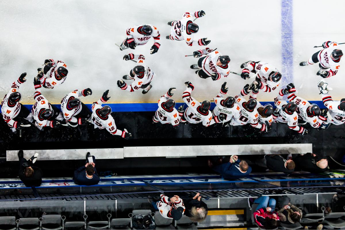 A photo taken from above, hockey players in white jerseys on the ice give fist bumps to teammates on the bench. Coaches in suits standing behind the bench jot down notes in small notebooks.