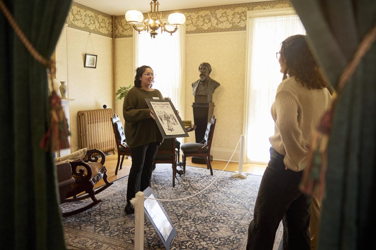 A tour guide standing in the back room of the Susan B. Anthony House with a tour group holding up an image of what the room originally looked like when Susan B. Anthony herself lived in the home.