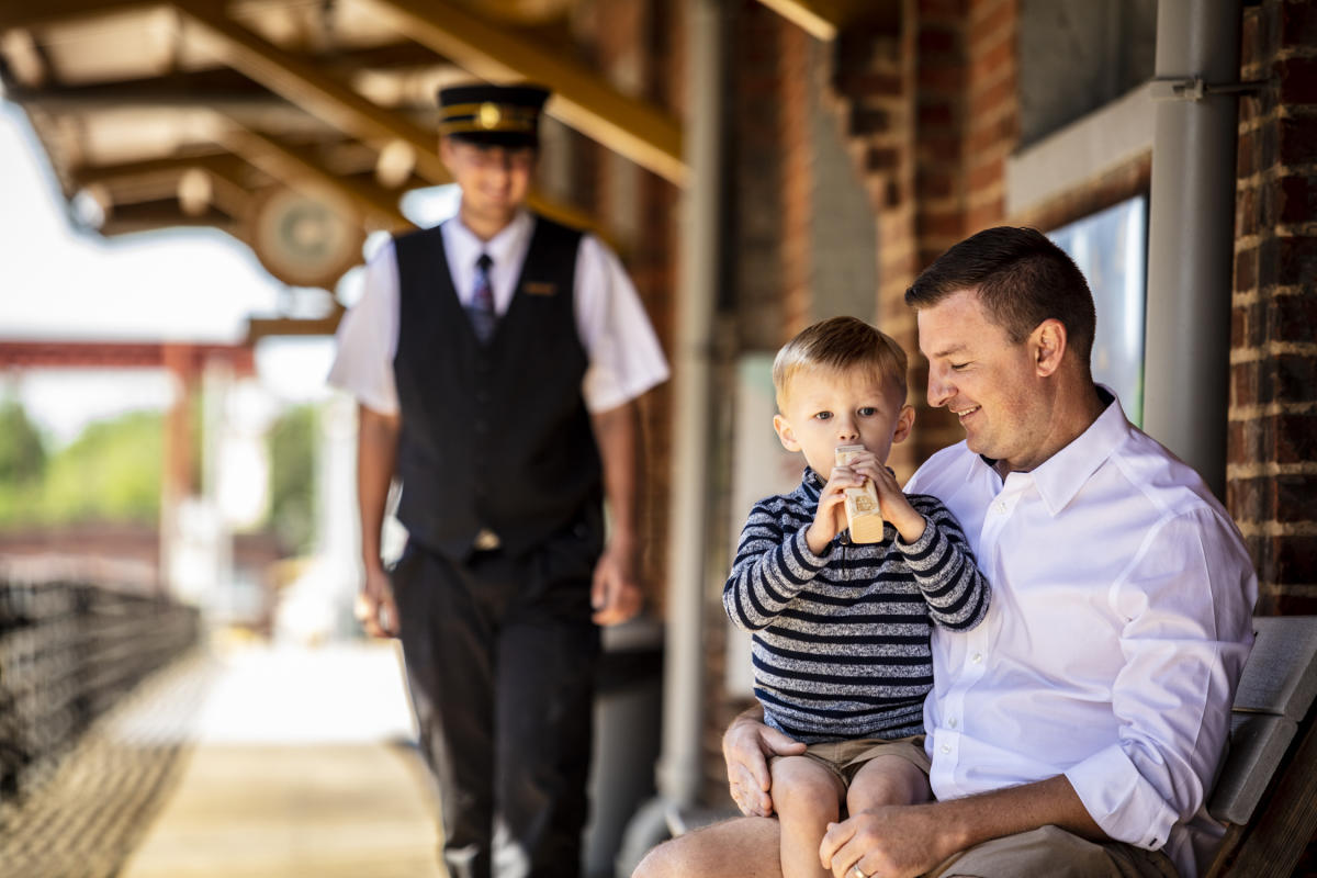 Dad and son on bench with train conductor in background