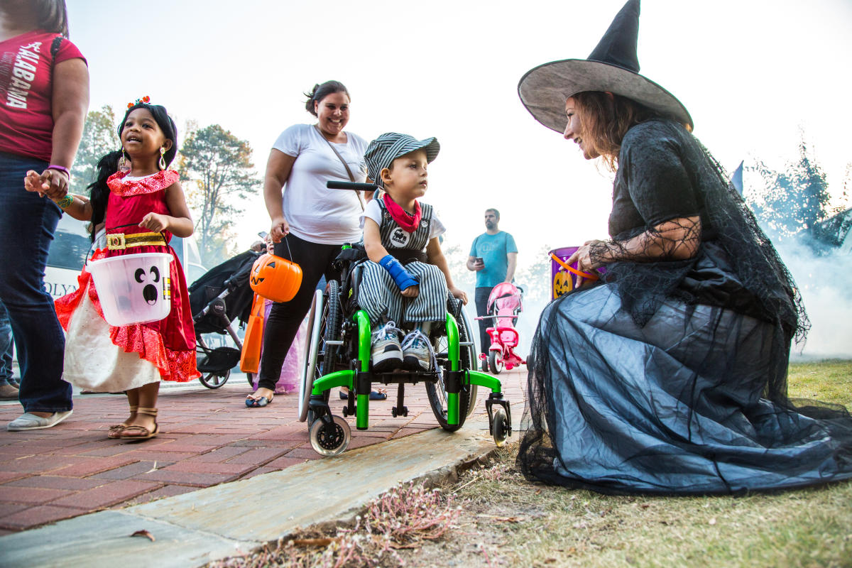 Adorable child in wheelchair with his guardian and Spooky Springs Witch character trick-or-treating.