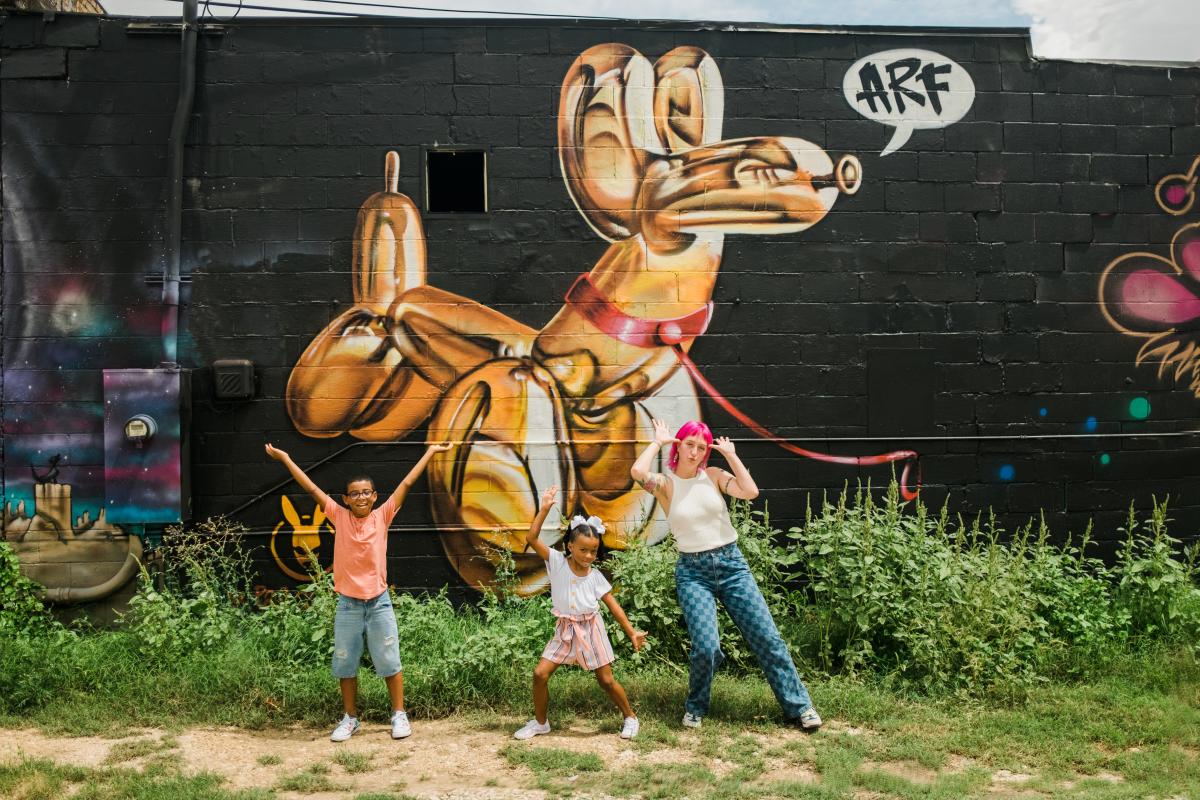 Woman and kids strike silly poses in front of a mural in Downtown San Marcos