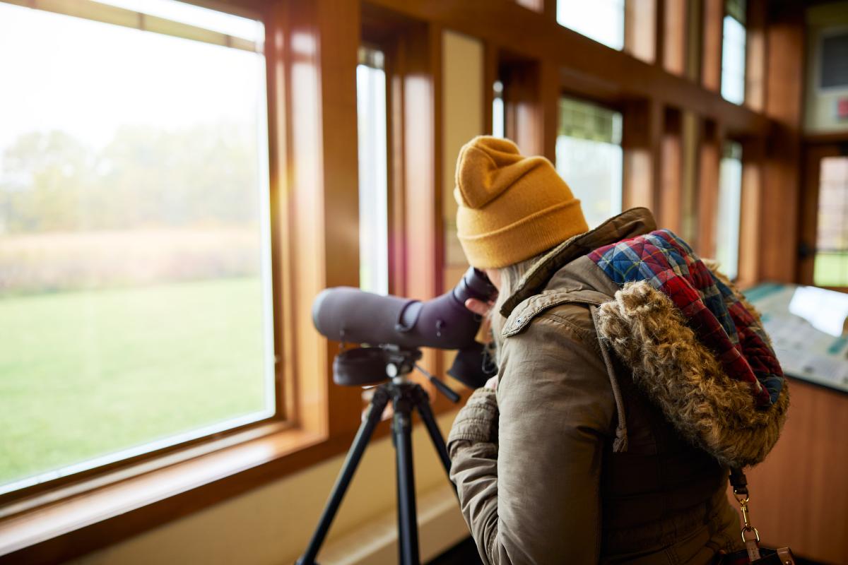 girl in yellow hat and parka looking through a telescope out a window