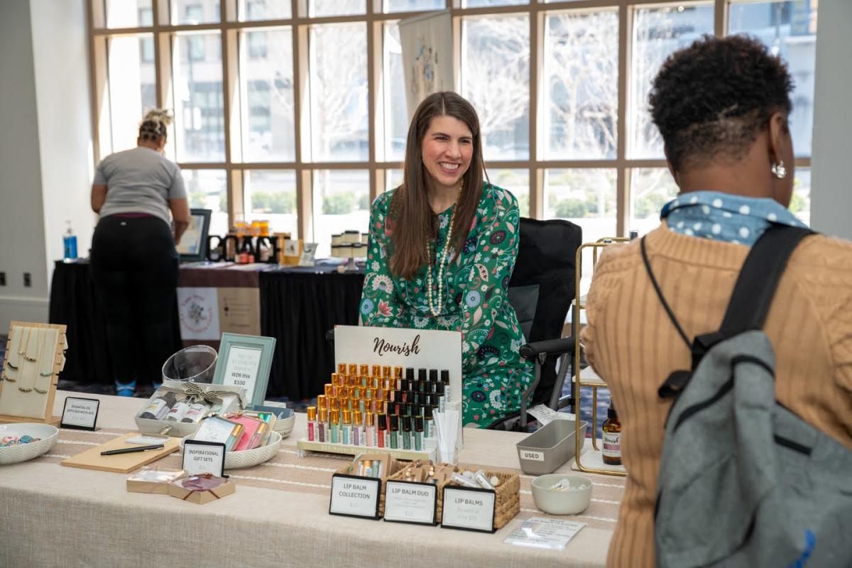 A woman smiles as patrons browse her selection of products at a market table.