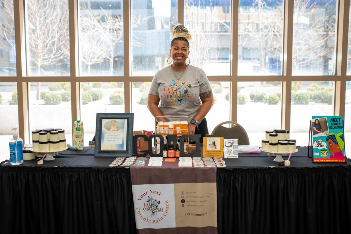 A woman stands behind a table of wellness products at a tradeshow.