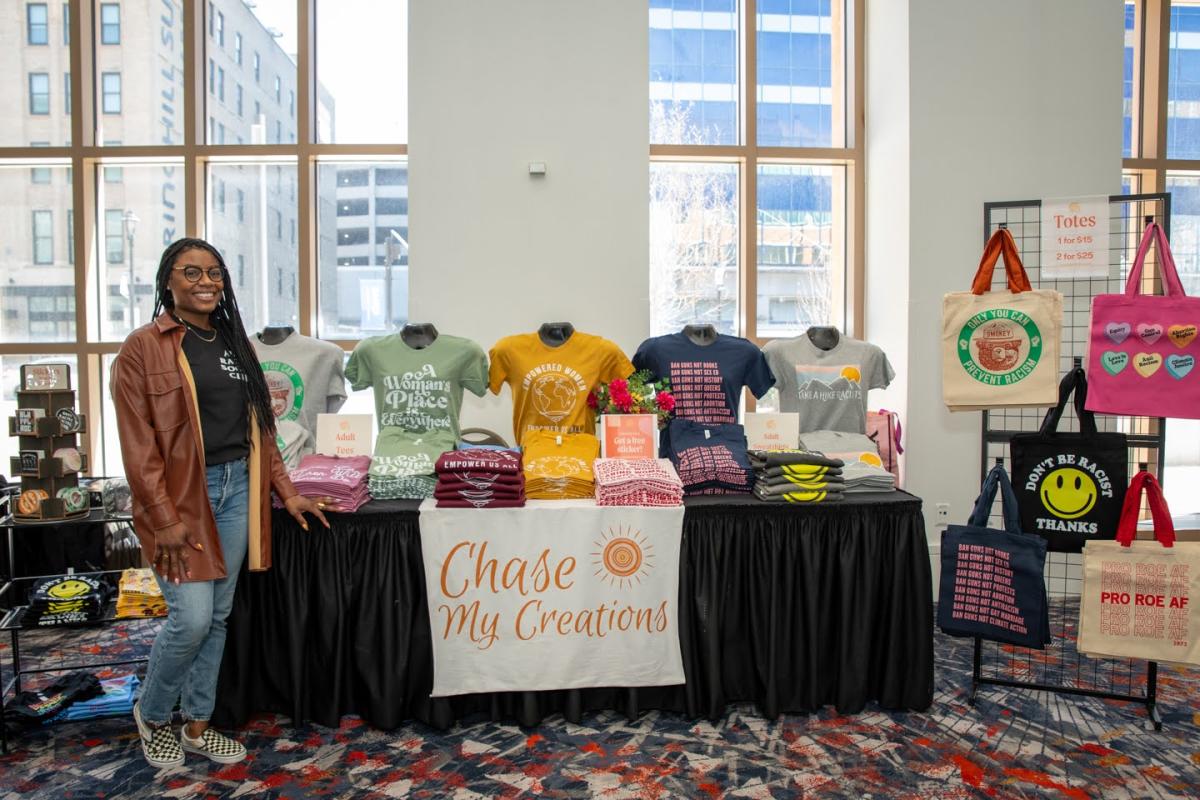 A woman posing in front of her marketplace table, consisting of custom designed t-shirts.