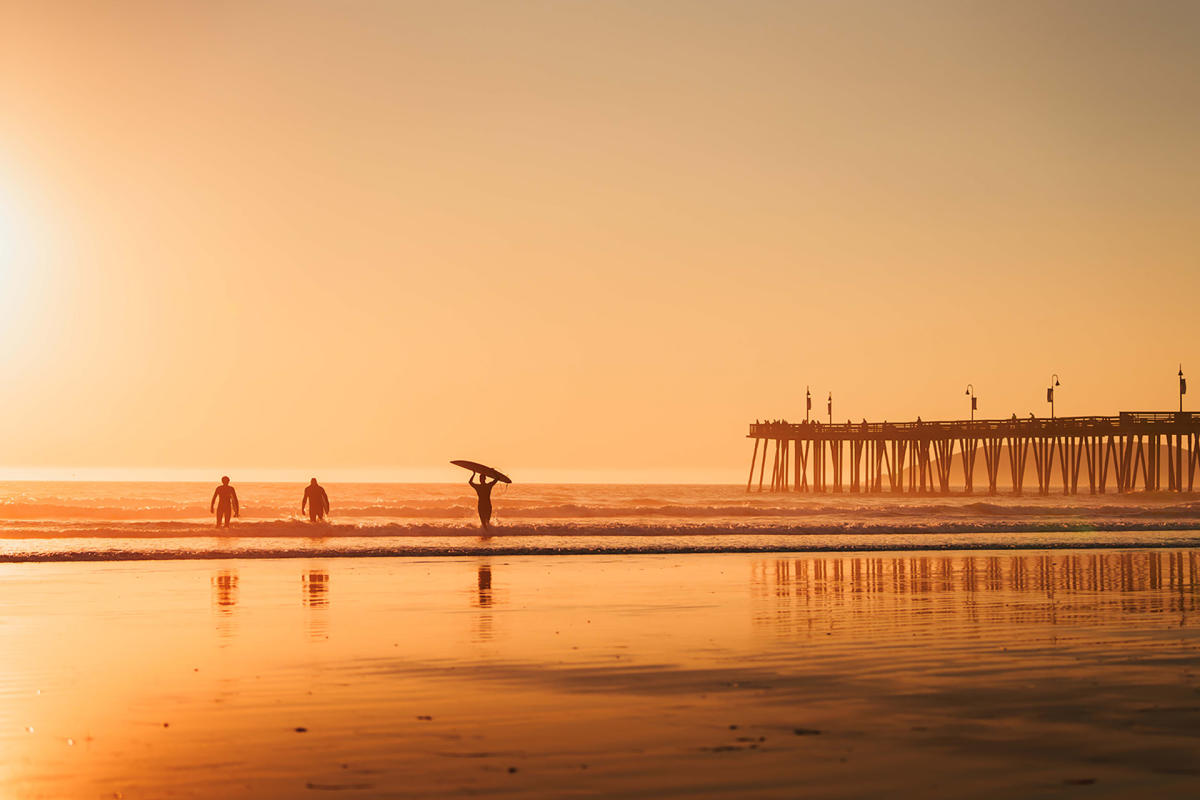 Three surfers carrying boards into the water next to a pier at sunset