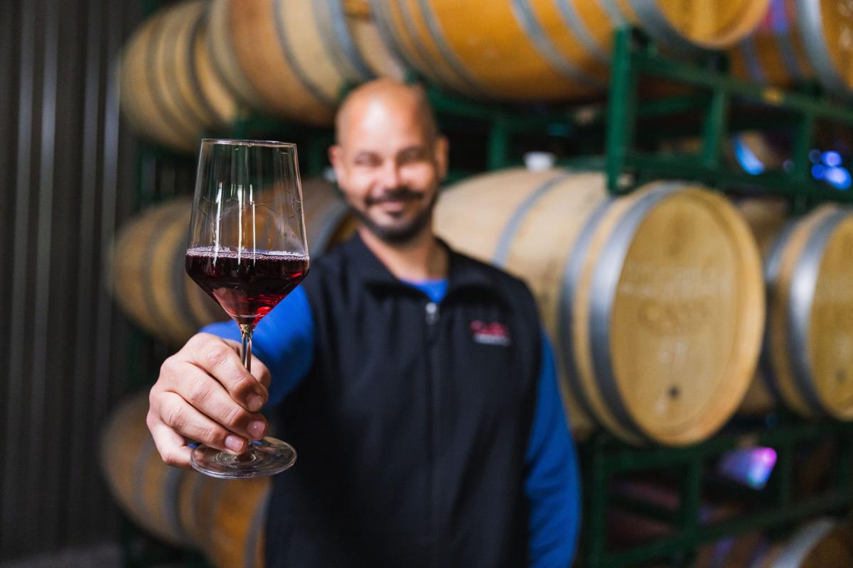 Man in soft focus in front of wine barrels holding a glass of red wine that is in focus