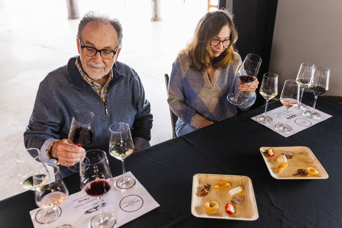 Two people sitting at table with multiple wine glasses and food platters, looking at wine