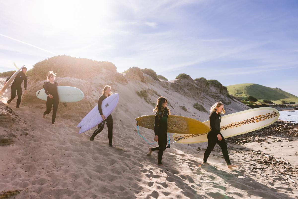 5 women in wetsuits carrying surfboards over a sand dune