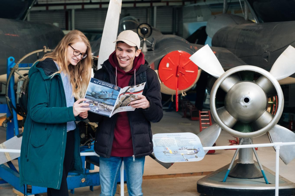 Two people looking at a leaflet and smiling stood in front old plane at Solent Sky musuem