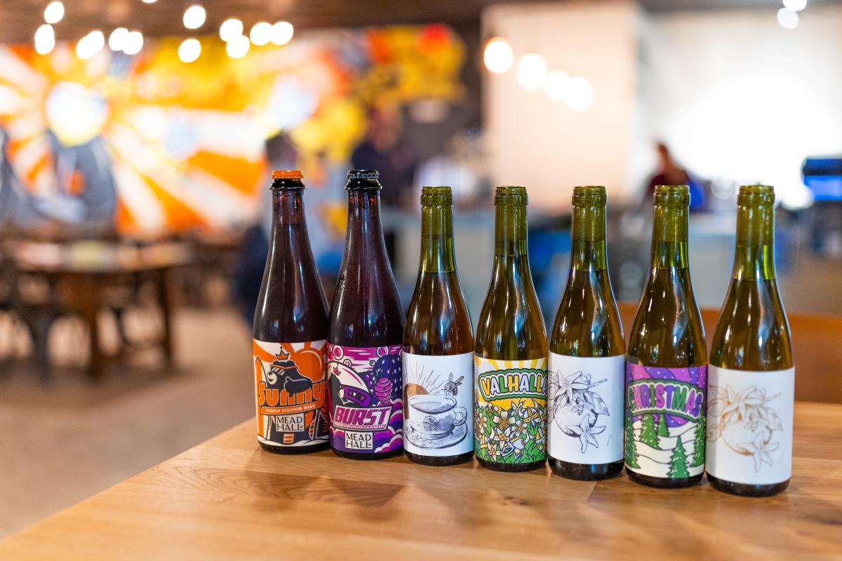 Lineup of colorful bottles of mead on table with tables and mural in background