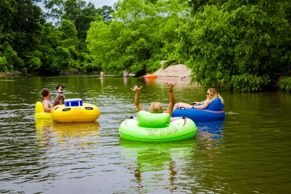Two women, a man and a dog sit atop brightly colored river tubes to float down Bogue Chitto River in Washington Parish, Louisiana, with swimmers in the distance. An extra tube holds a blue cooler. The area is surrounded by lush, green trees and bushes.