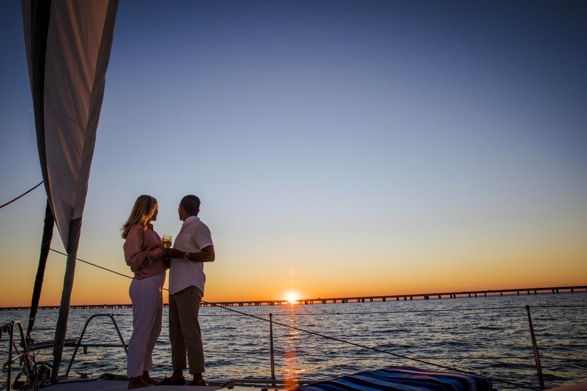 A man and a woman stand on a sailboat watching the sun set over the Lake Pontchartrain Causeway Bridge.