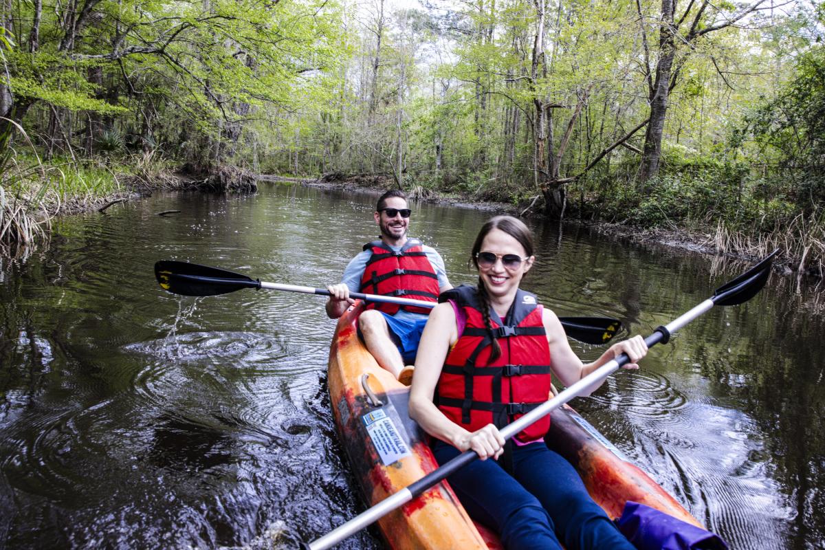 Two people in red life jackets kayak on the calm, tree-lined Bogue Falaya River. Lush greenery surrounds the serene scene, reflecting in the water.