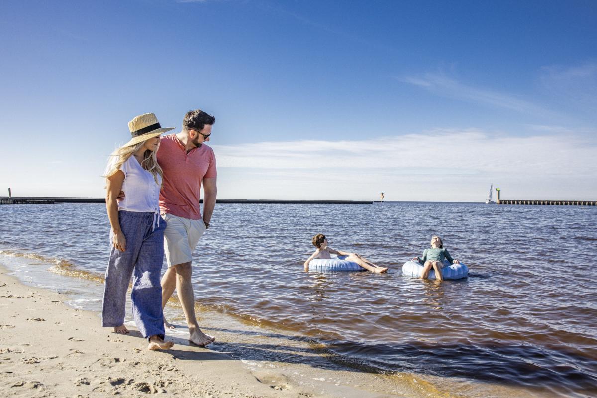 A couple strolls along the sandy beach at Fontainebleau State Park while two children relax on inner tubes in the calm waters of Lake Pontchartrain beneath a clear summer sky.