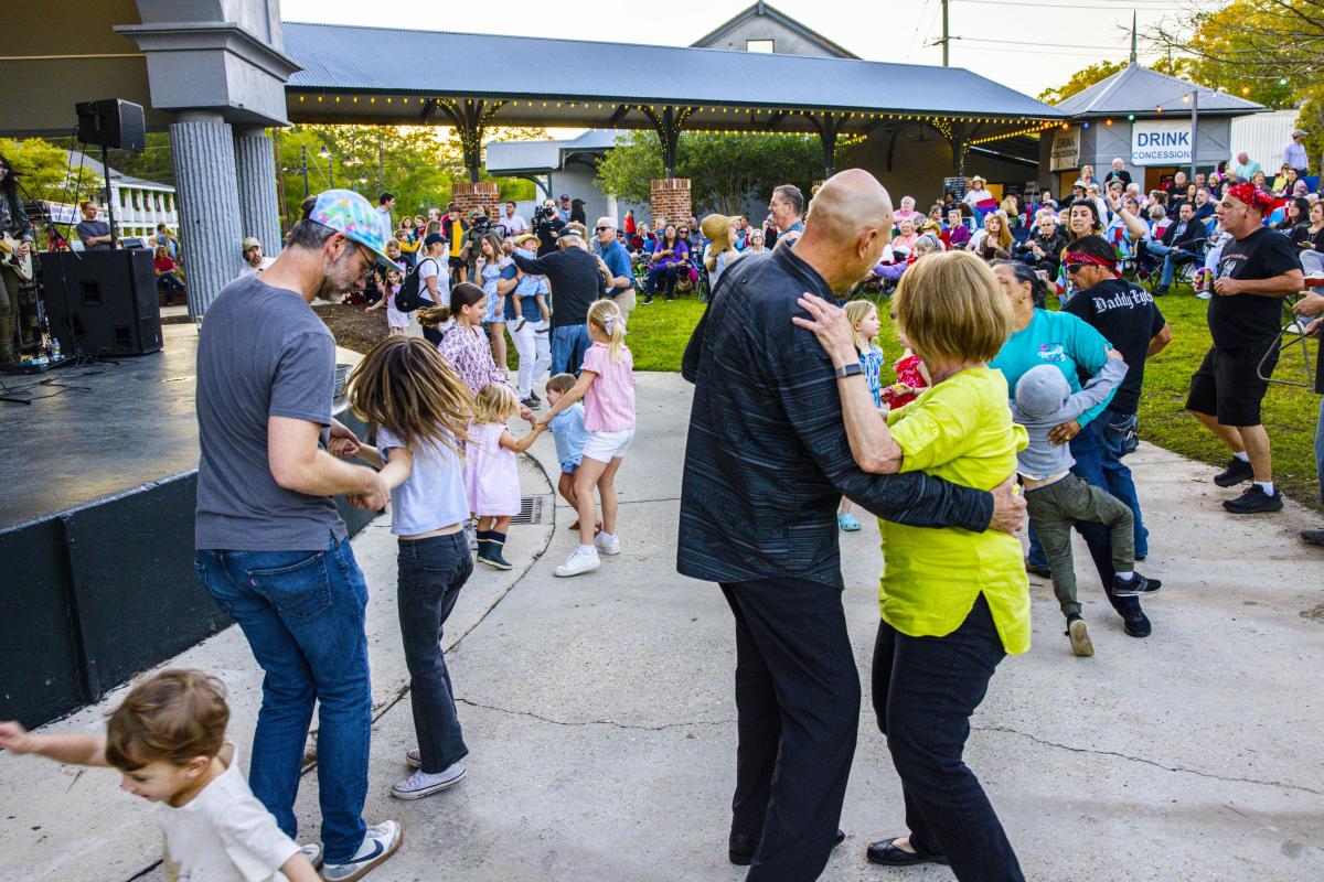 People of all ages dance near the stage during a Mandeville Live! free Friday concert at the Mandeville Trailhead, with a crowd gathered under the pavilion and on the lawn.