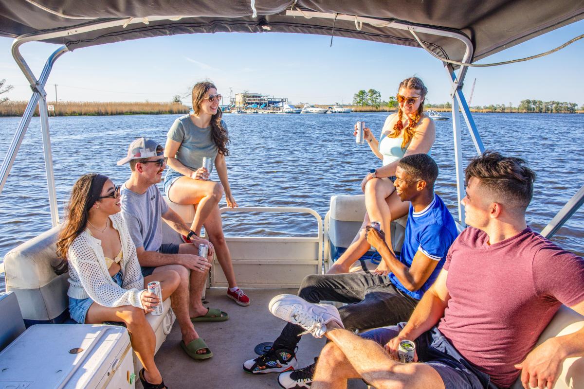 Six young people are seated in a canopied pontoon boat, talking and drinking, with the Tchefuncte River behind.