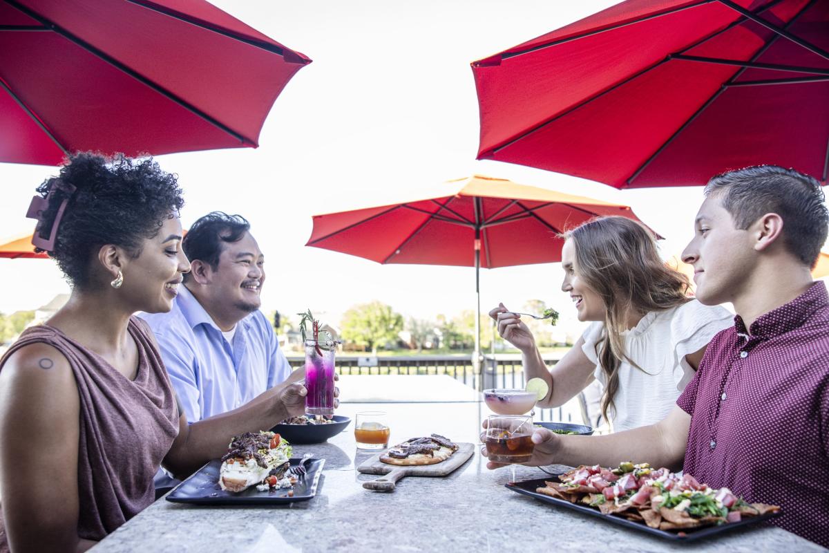 Two couples sit at an outdoor patio under red umbrellas eating seafood.