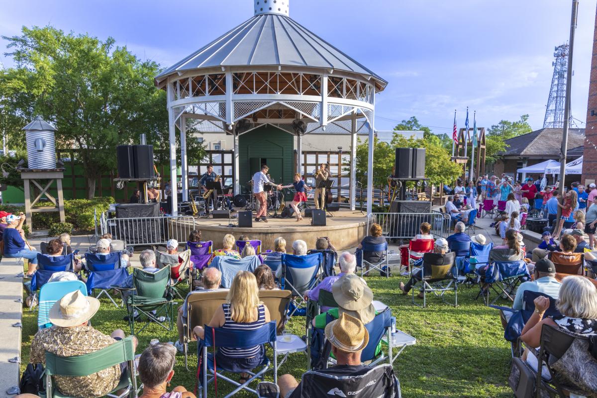 A crowd gathers in lawn chairs around the pavilion at the Covington Trailhead as a live band performs onstage during an outdoor concert.