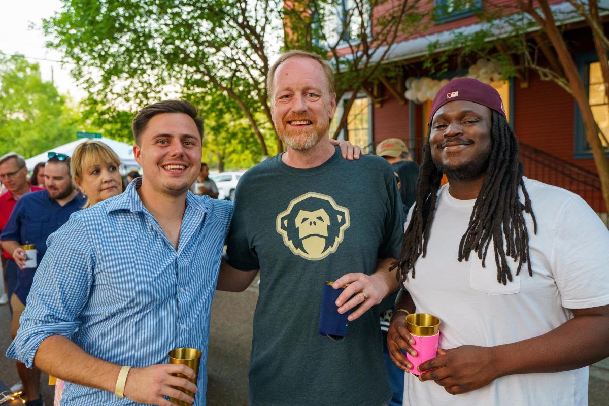 Three men stand together smiling and holding drinks during the Girod Street Stroll in Mandeville, with a crowd and historic buildings in the background.