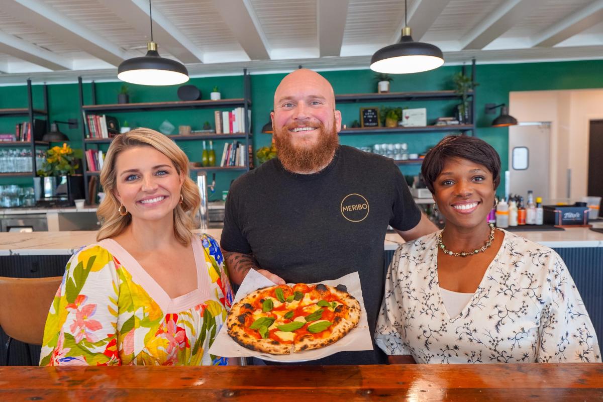Katie Guasco and Zondra White Jones smile alongside Chef Gavin Jobe of Meribo, who holds a wood-fired Margherita pizza inside the restaurant's bright, modern dining room.