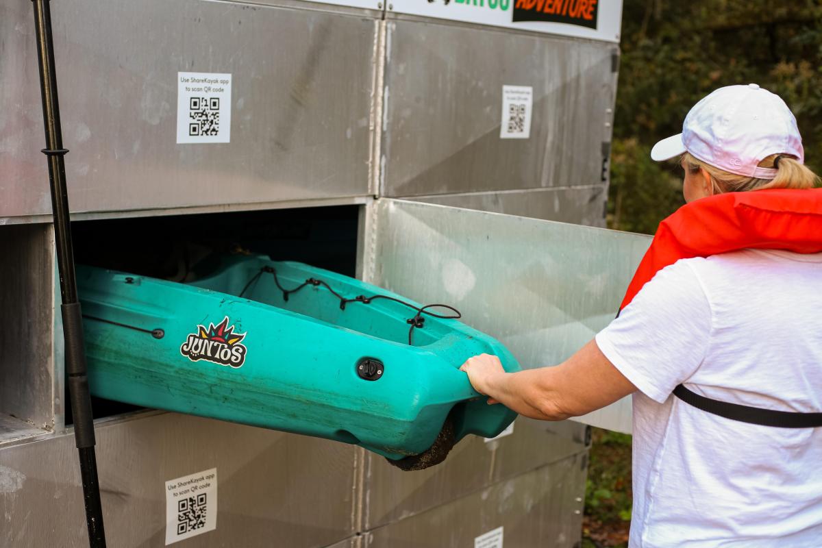 A person in a life jacket retrieves a turquoise kayak from a storage unit, surrounded by QR codes and nature.