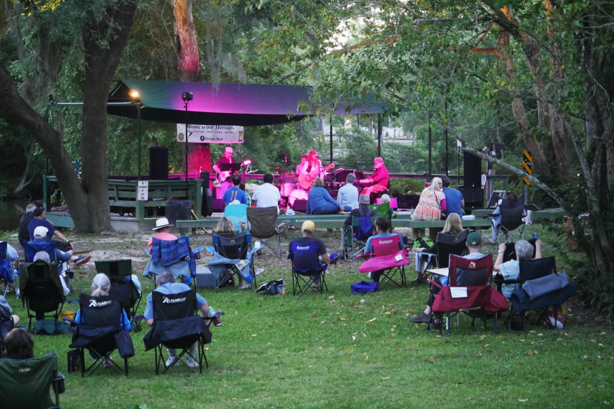 Audience members sit in lawn chairs beneath the trees during a Sunset at the Landing concert in Covington, as musicians perform on the outdoor stage at dusk.