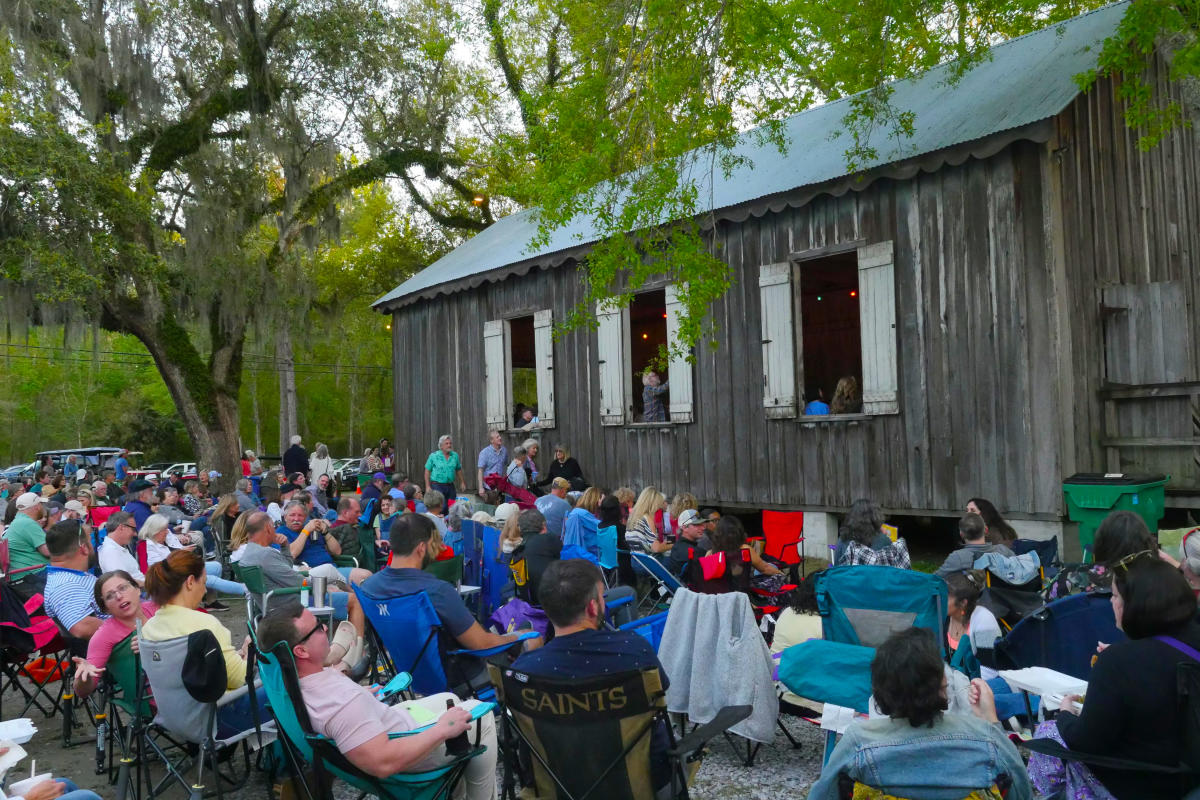 Concertgoers relax in folding chairs outside the historic Dew Drop Jazz Hall in Mandeville, listening through the open windows beneath moss-draped live oaks.