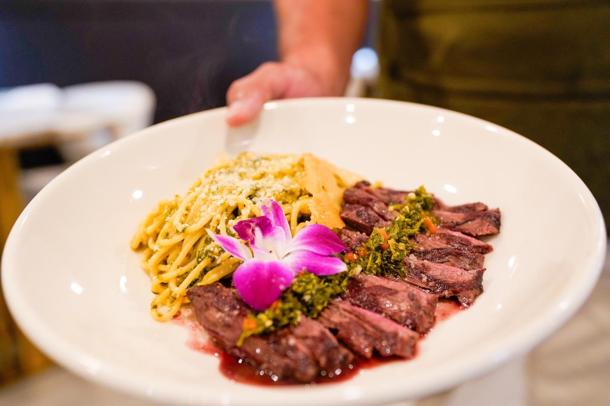 A plate of sliced meat and pasta with a hot pink edible flower on top