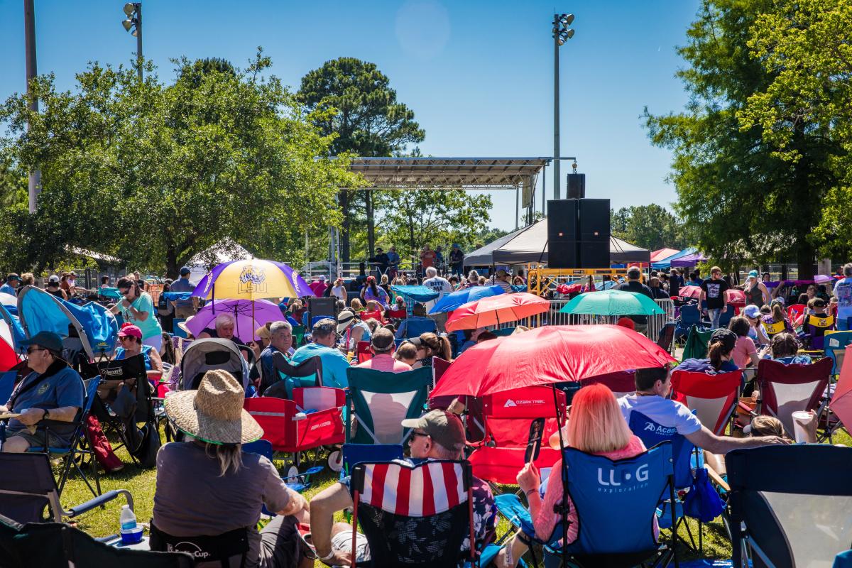 A large crowd sits in lawn chairs and under colorful umbrellas facing a stage during the Slidell All-You-Can-Eat Crawfish Cook-off on a sunny day.