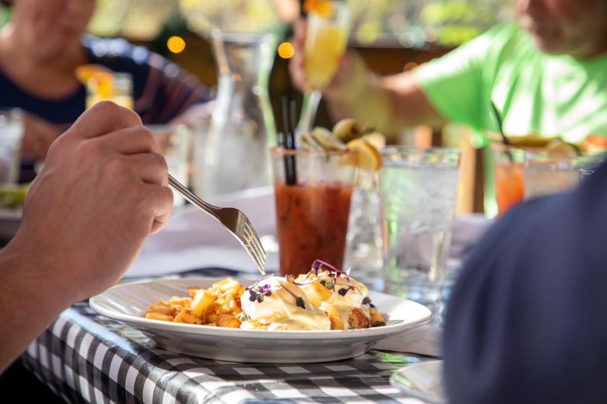 Indistinct persons sit around a checkered cloth-covered patio table laid with plates and glasses, eating brunch foods and drinking Bloody Marys and mimosas.