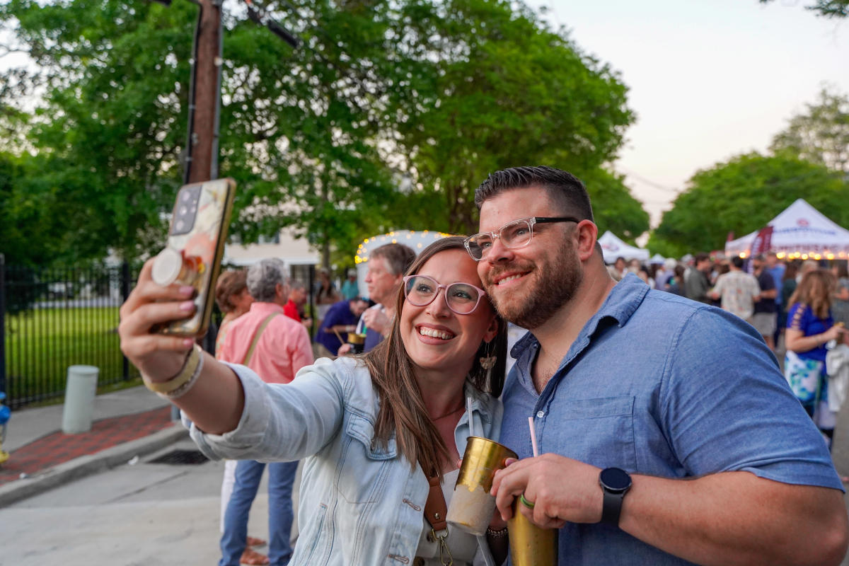 A couple smiles and takes a selfie while holding drinks during the Girod Street Stroll in Mandeville, with a lively crowd and vendor tents in the background.