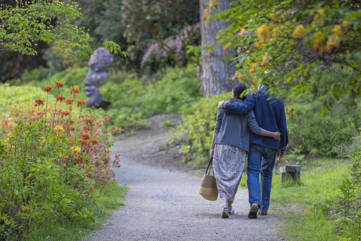 A couple walk along a path surrounded by flowers towards a large sculpture of a face in Leonardslee gardens, Sussex