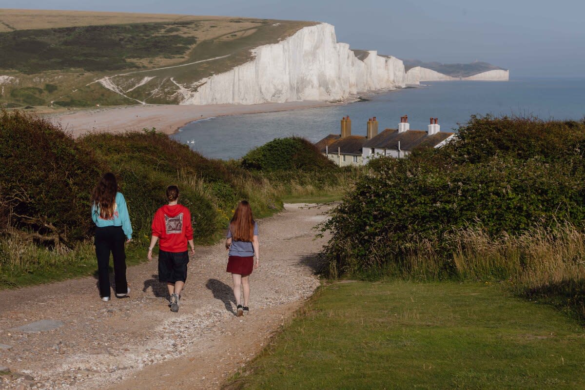 Looking towards Seven Sisters white chalk cliffs  from Seaford Head, three females are walking down a path away from the camera