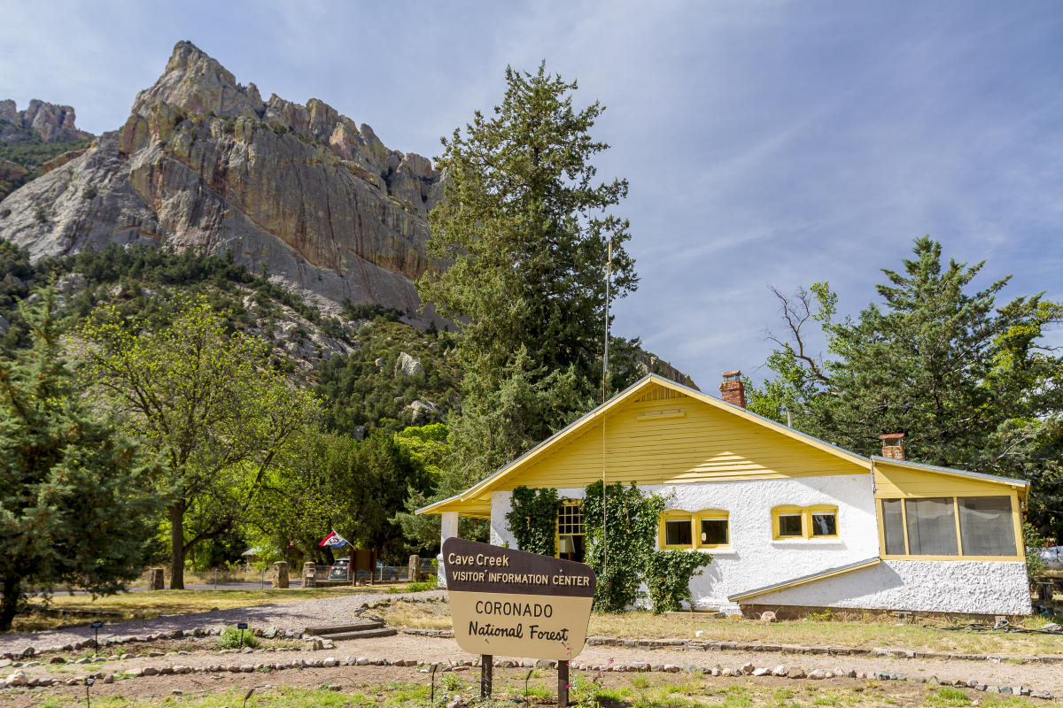 small white house with yellow roof and shutters next to a large mountain/rock and trees. In front a sign stands that says "Cave Creek Visitor Information Center, Coronado National Forest