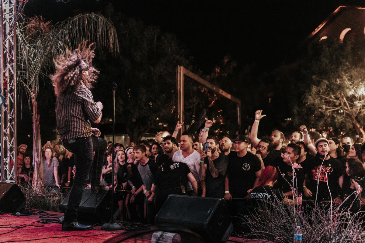 person thrashing their long hair while playing guitar on an outdoor stage in front of a large crowd of people